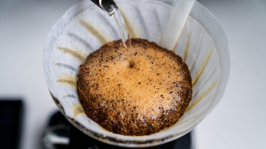 A close-up shot captures freshly ground coffee in a white paper filter, showing the distinctive dome-like blooming ef...