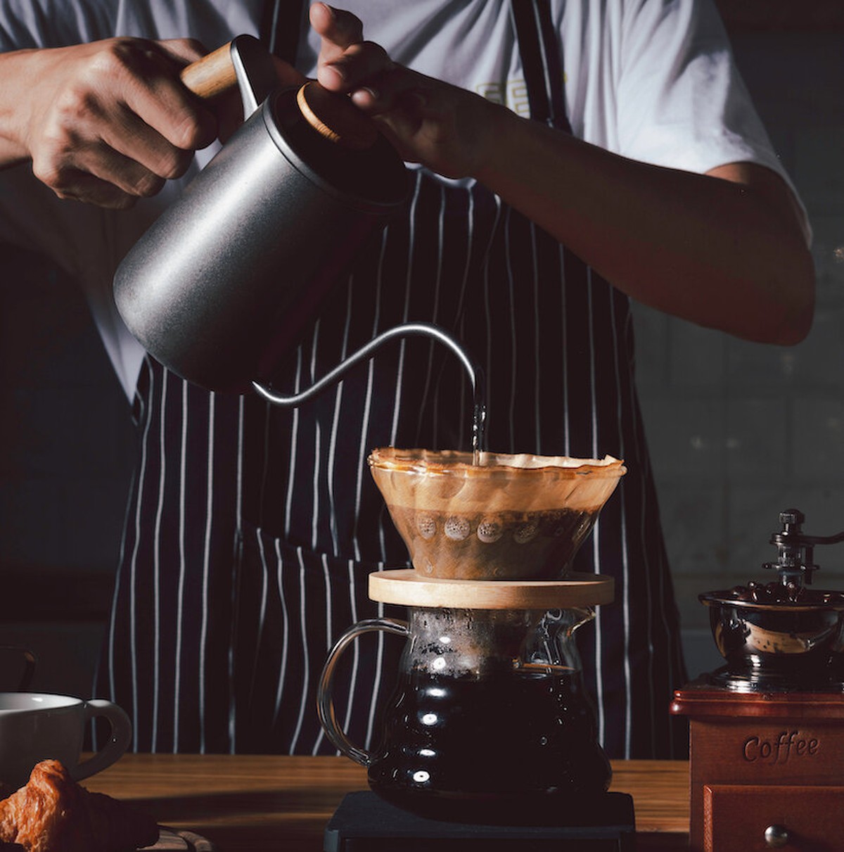 A barista in a striped apron performs a precise pour-over using a gooseneck kettle over a V60 dripper, with a manual...